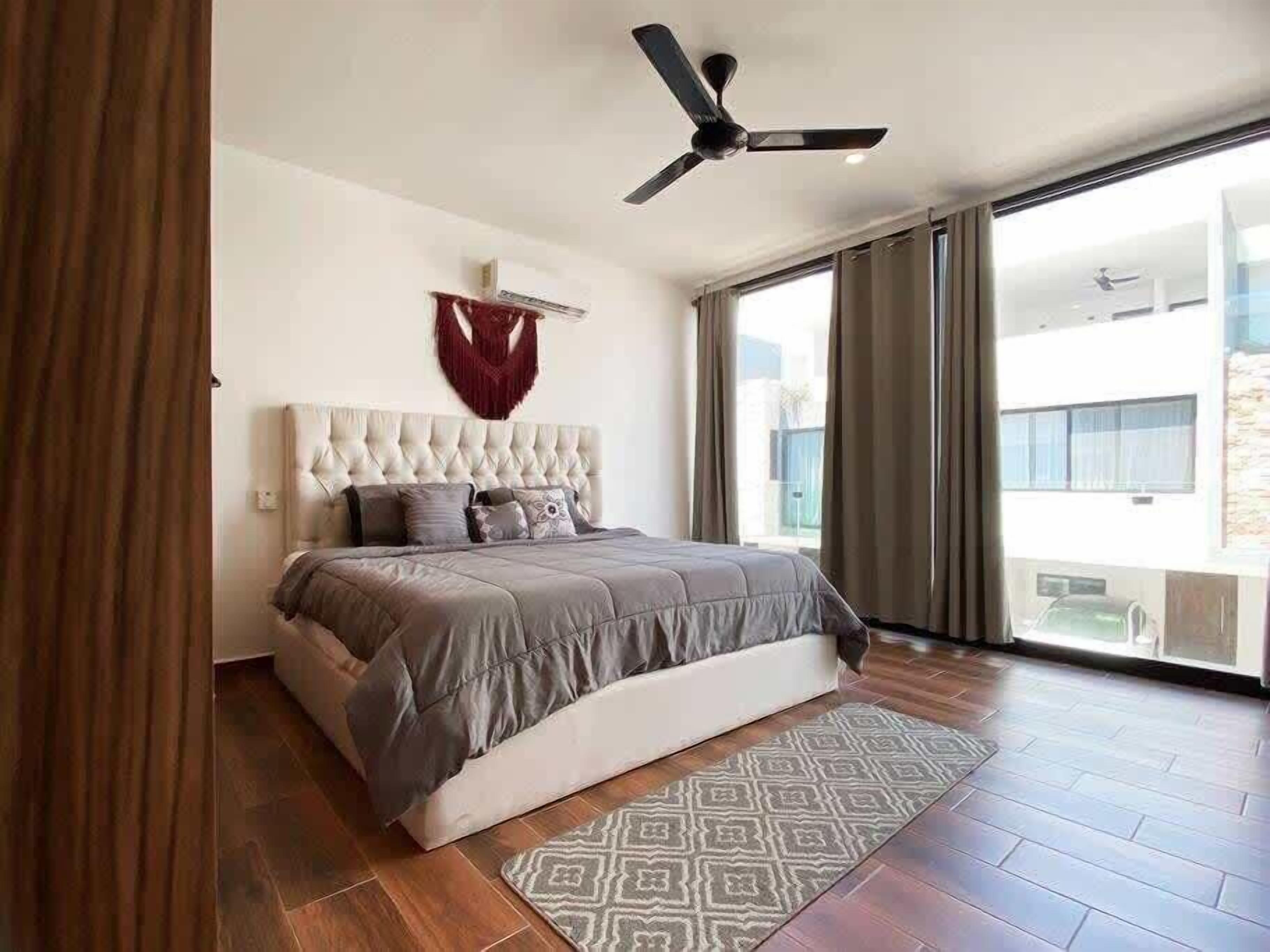 A bedroom with a tufted headboard, a ceiling fan, a patterned rug, and a view of a building through large windows.