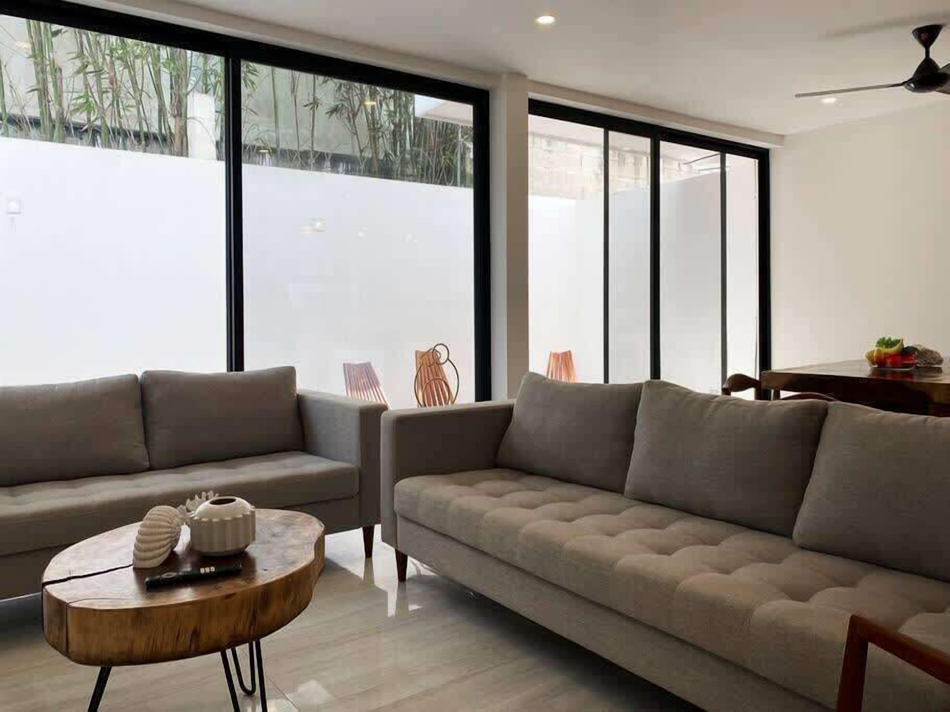 A modern living room with a sofa, a round wooden coffee table, and large windows with a view of bamboo outside.