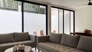 A modern living room with a sofa, a round wooden coffee table, and large windows with a view of bamboo outside. - 4BR House · Sleeps 12 · Shared Pool · Playa (Playa del Carmen)