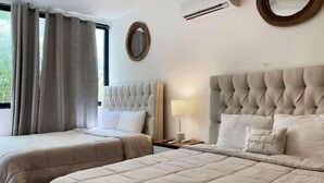 A hotel room with two beds, a round mirror, an air conditioning unit, and a window with curtains. - 4BR House · Sleeps 12 · Shared Pool · Playa (Playa del Carmen)