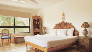 A bedroom with a wooden bed, a nightstand, a bookshelf, a chair, and a window with a view of palm trees. - 2BR Cancun Hotel Zone: Pool, Walk to the Beach (Cancún)