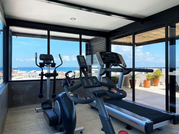 A gym with treadmills and elliptical machines, large windows, and a view of the city. - 2BR Apt · Rooftop Pool & Gym · Near 5th Ave (Playa del Carmen)