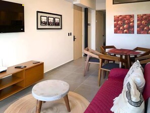 A living room with a TV, a dining table with chairs, a red sofa, and a small round table with a stool. - 2BR Apt · Rooftop Pool & Gym · Near 5th Ave (Playa del Carmen)