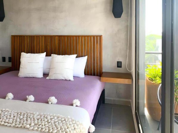 A bedroom with a wooden headboard, a purple bedspread, white pillows, and a window with a view of greenery. - 2BR Apt · Rooftop Pool & Gym · Near 5th Ave (Playa del Carmen)