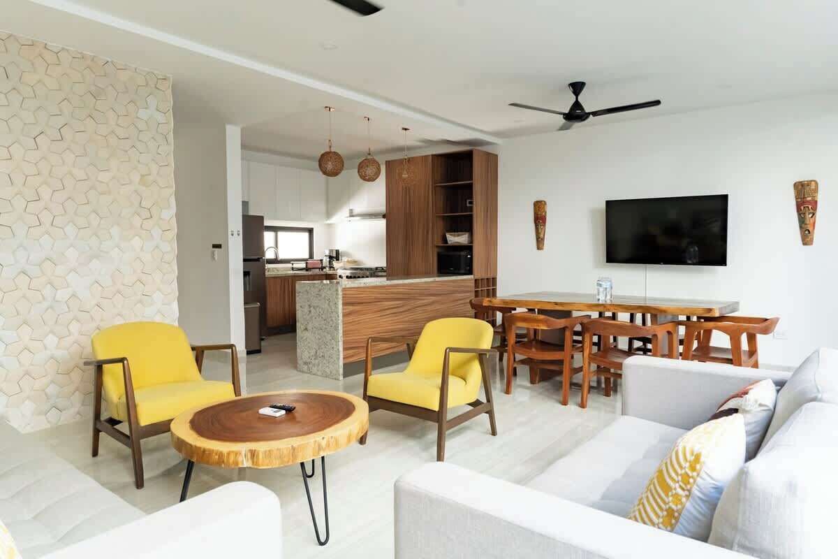 Un salon moderne avec un canapé, des fauteuils jaunes, une table basse ronde en bois, un coin repas avec une table et des chaises, une cuisine en arrière-plan et un téléviseur à écran plat fixé au mur.