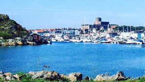 Un pueblo costero con un puerto deportivo lleno de barcos, una imponente edificación similar a un castillo y terreno rocoso.