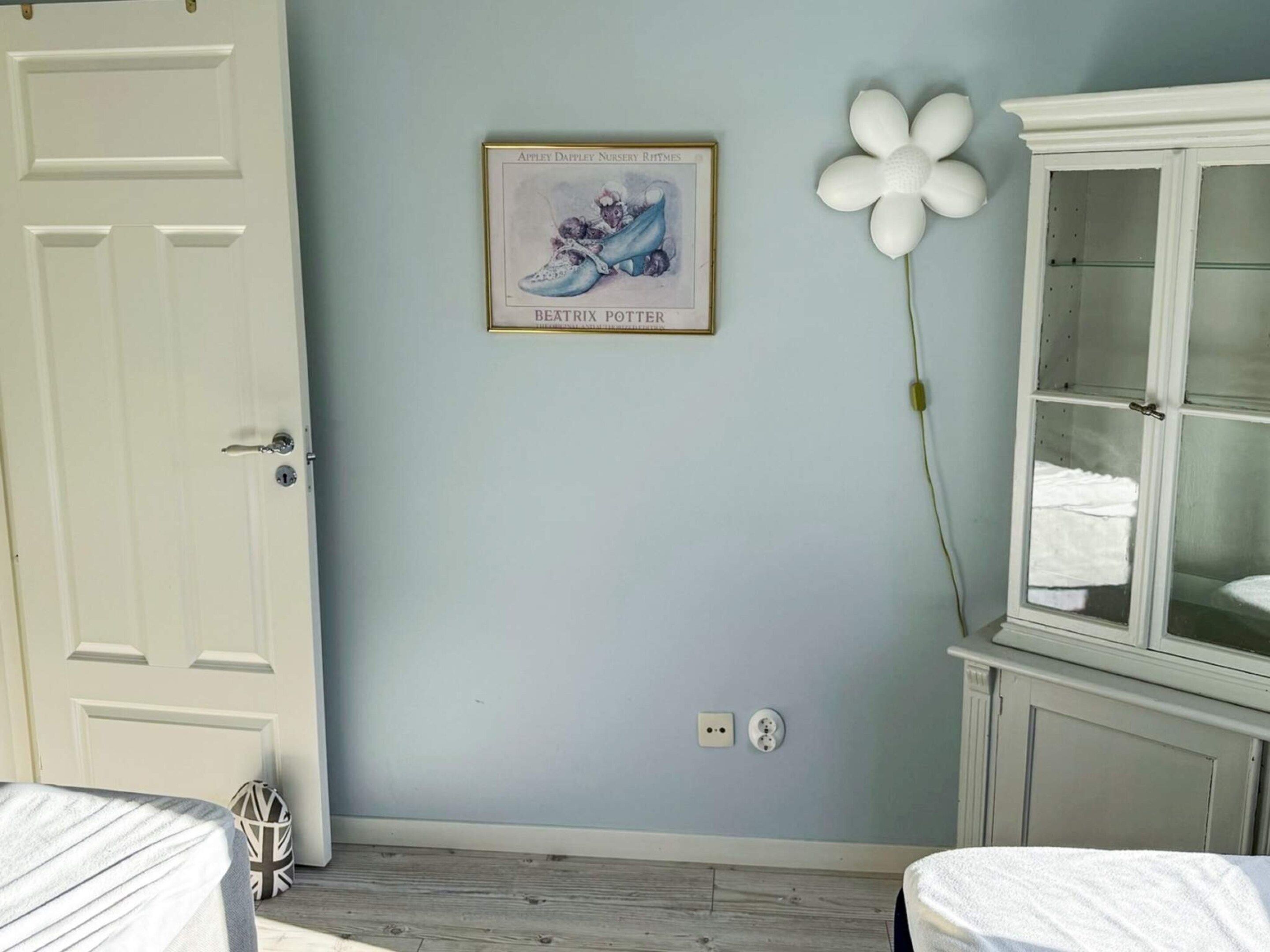 A room with a white door, a framed nursery rhyme poster, a white flower decoration, a cabinet with glass doors, and a bed with a white cover.
