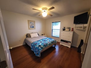 A bedroom with a bed, a dresser, a ceiling fan, a window, and a wall-mounted TV. - Central Florida Pool Home (DeBary)