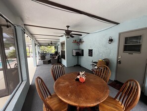 A covered patio with a round wooden table and chairs, ceiling fans, and a TV on a stand. - Central Florida Pool Home (DeBary)