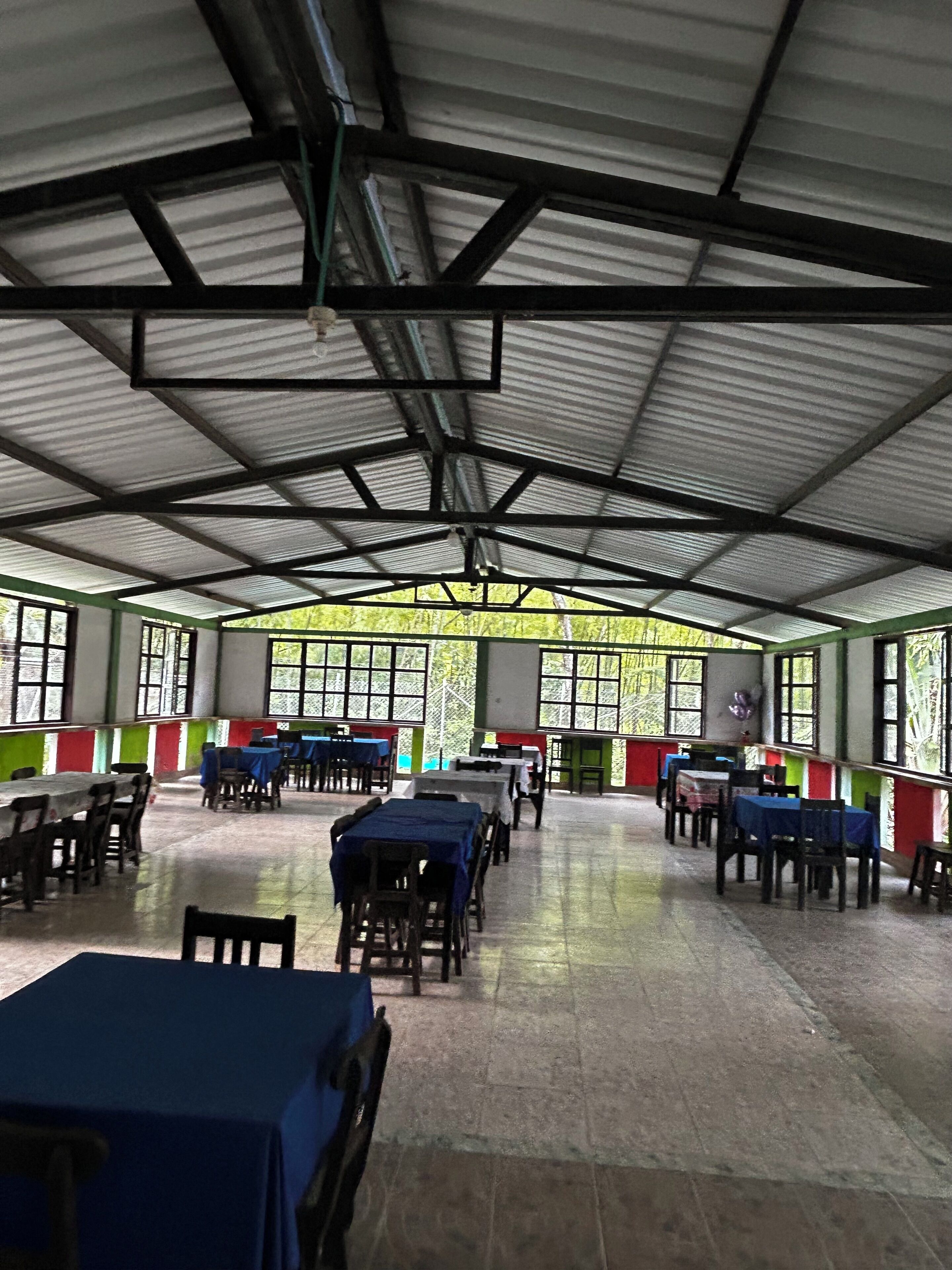 Un área de comedor techada con mesas y sillas, ventanas amplias y estructura de techo metálica.