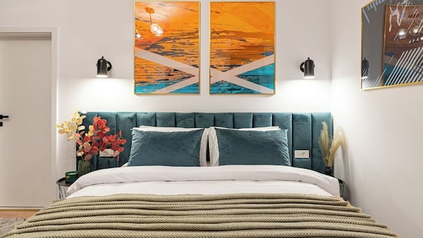 A modern bedroom with a teal upholstered headboard, a large bed with white bedding, two wall-mounted paintings, and a nightstand with a vase of flowers. - Central Square Haven B (București)