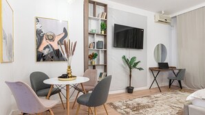 Een moderne woonkamer met een ronde eettafel, grijze stoelen, een boekenkast, een flatscreen televisie, een bureau met een laptop, een potplant en een grote spiegel.