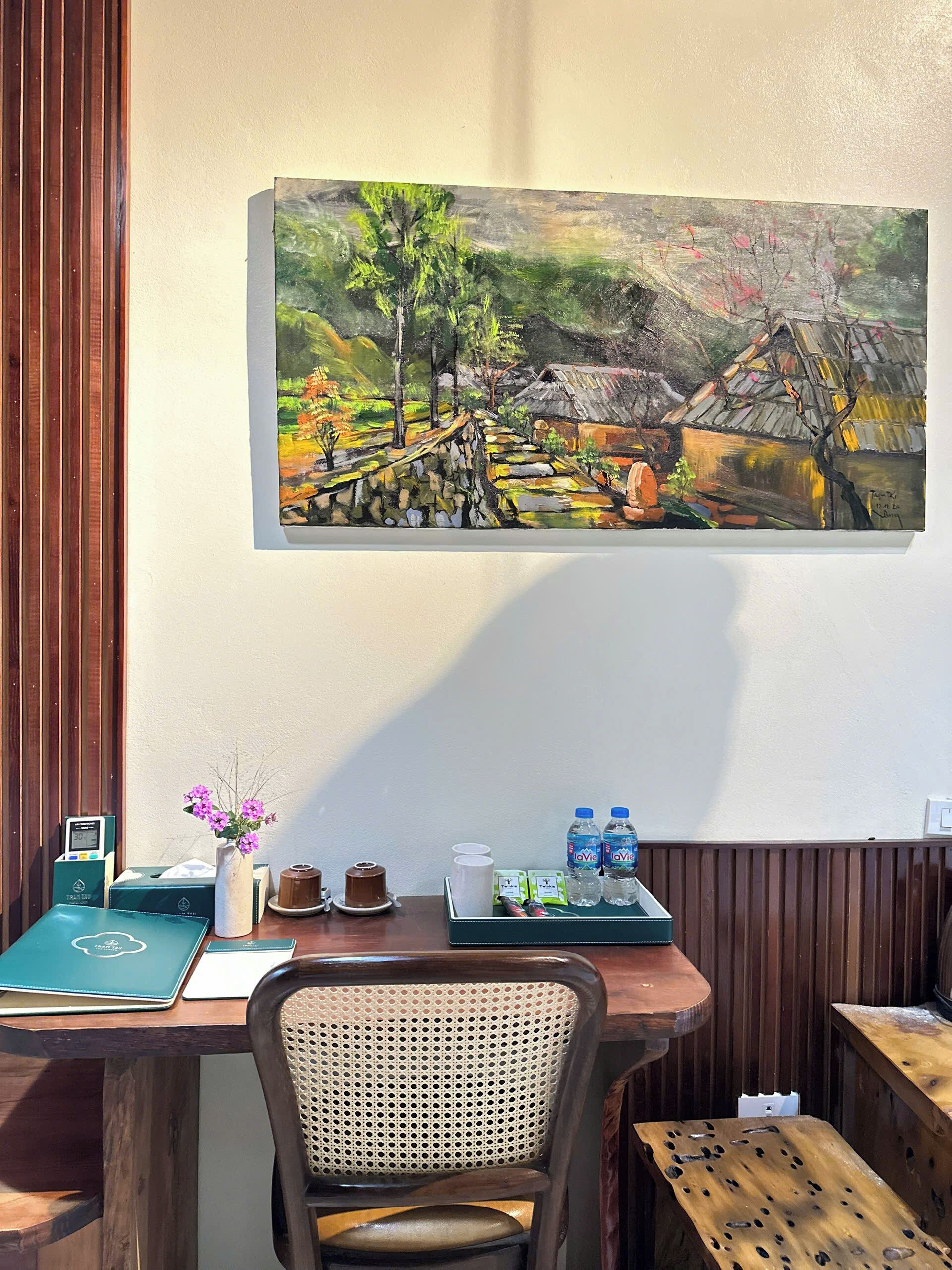 A wooden table with a painting, a chair, a vase with flowers, and a tray with water bottles and a cup.