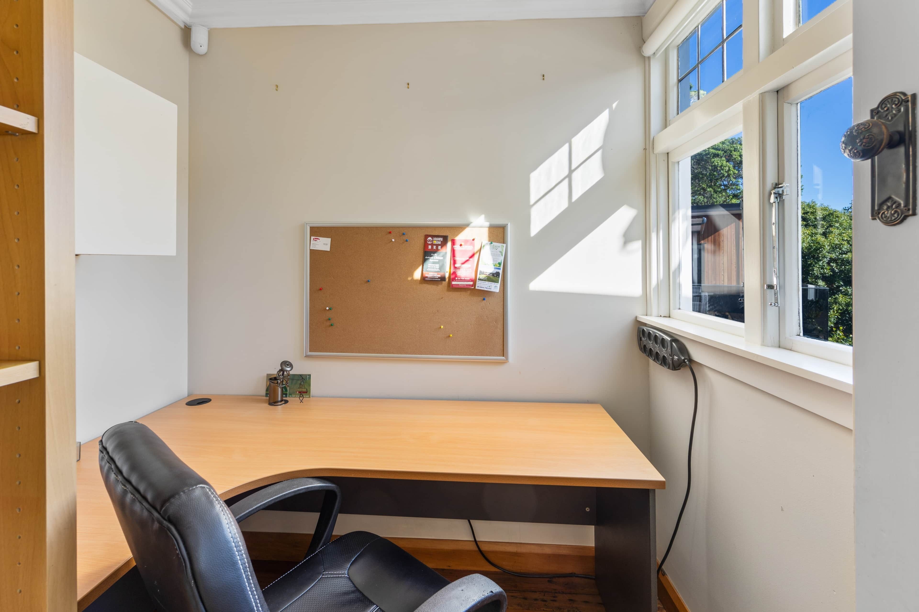 A small office with a wooden desk, a black leather chair, a corkboard with notes and a calendar, and a window with blinds.