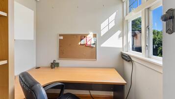 A small office with a wooden desk, a black leather chair, a corkboard with notes and a calendar, and a window with blinds.