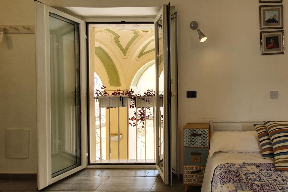 Un dormitorio con cama, mesita de noche, lámpara de pared y puerta que da a un balcón con vistas a un edificio con arcos.