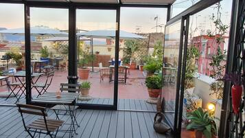 Een overdekt terras met tafels en stoelen, potplanten en een uitzicht op de stadsachtergrond.