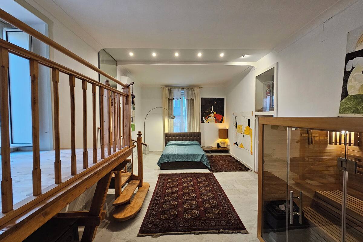 Un dormitorio moderno con una escalera de madera, una cama con colcha estampada, una alfombra y un aparador de vidrio.