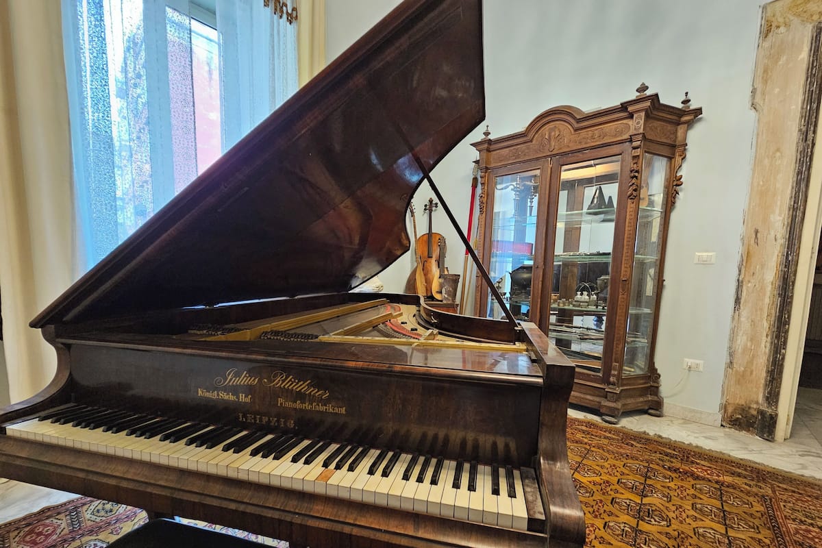 Un piano de cola en una habitación con alfombra estampada, un mueble de madera con puertas de cristal y una ventana con cortinas decoradas.