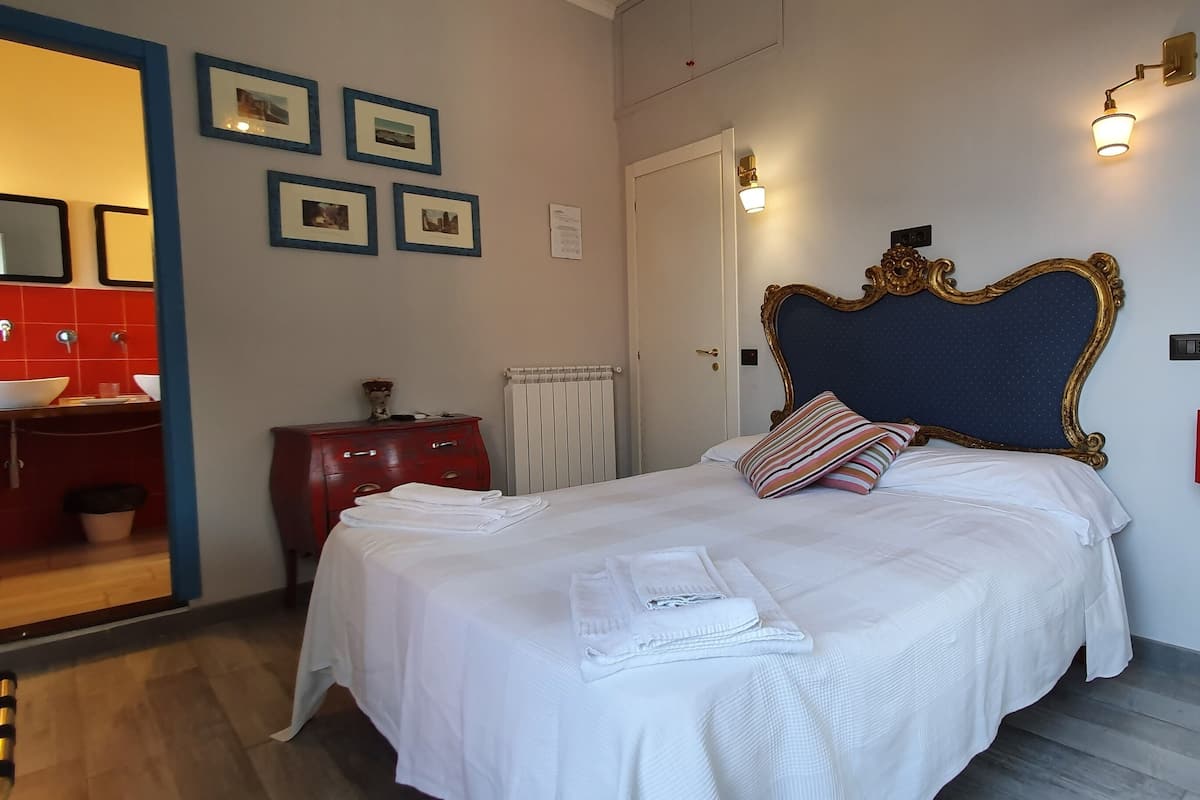 Habitación de hotel con una cama, un aparador rojo, un radiador blanco y un baño visible a través de una puerta abierta.