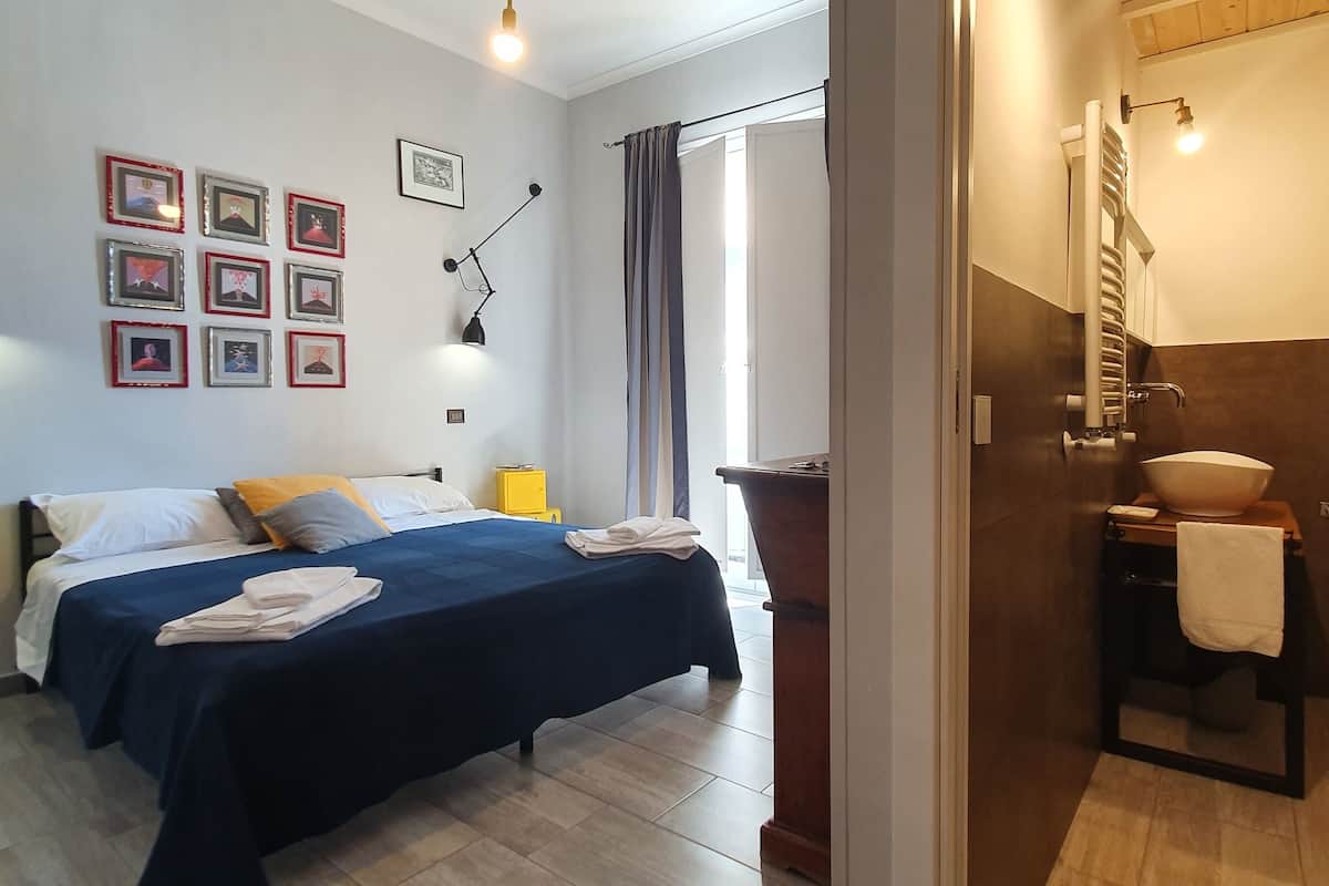 Habitación de hotel con cama, lámparas de noche, una mesita con una toalla, un espejo y un baño con inodoro y lavamanos.