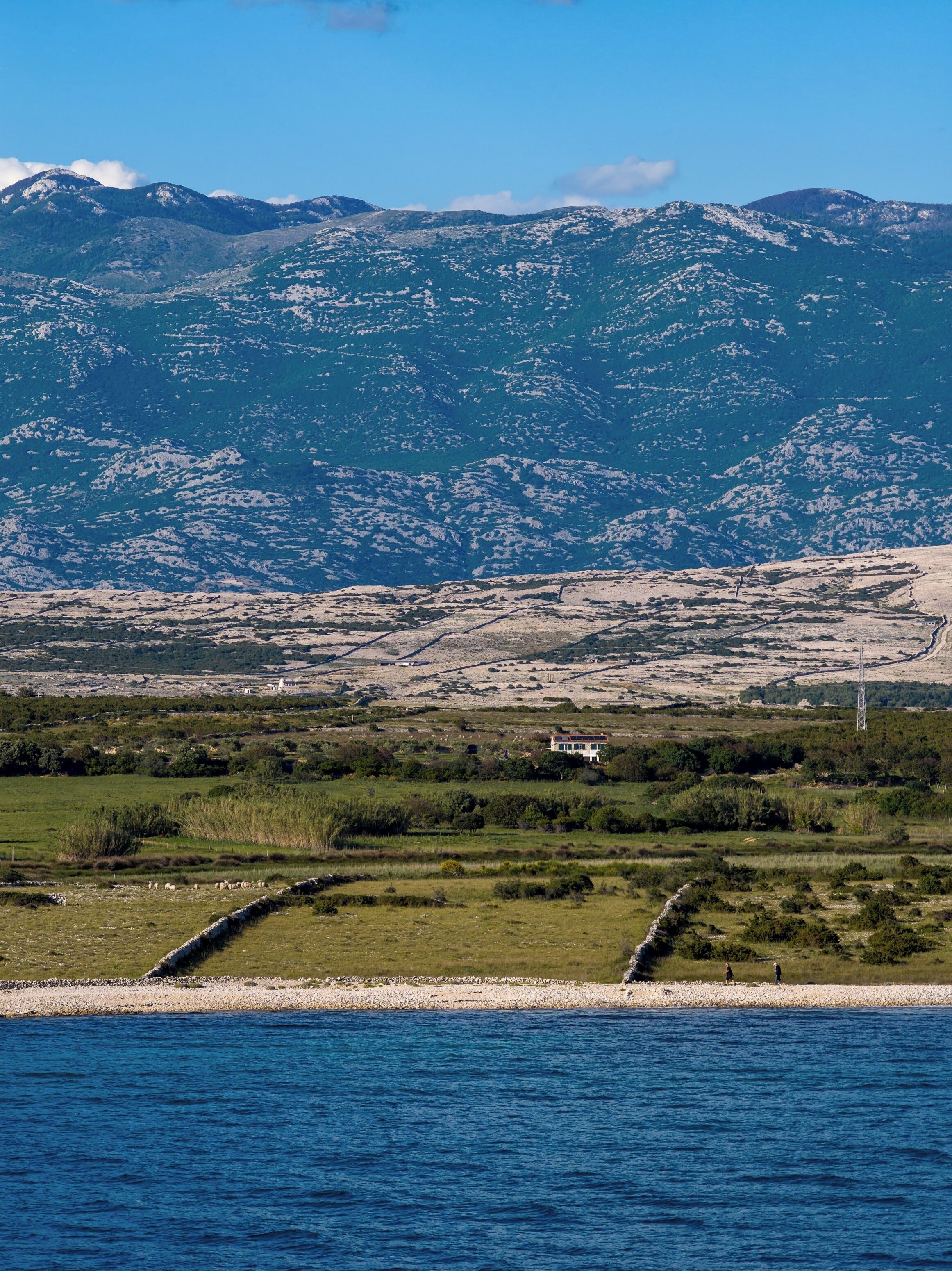 A coastal landscape with mountains, a body of water, and a house with a stone wall in the foreground.