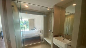 Una camera d'albergo moderna con un divisorio in vetro che separa la zona notte dal bagno. La stanza presenta un letto grande, un lavandino bianco e uno specchio sopra il lavandino.