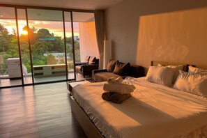 A hotel room with a large bed, a sofa, and a balcony with a view of the sunset.