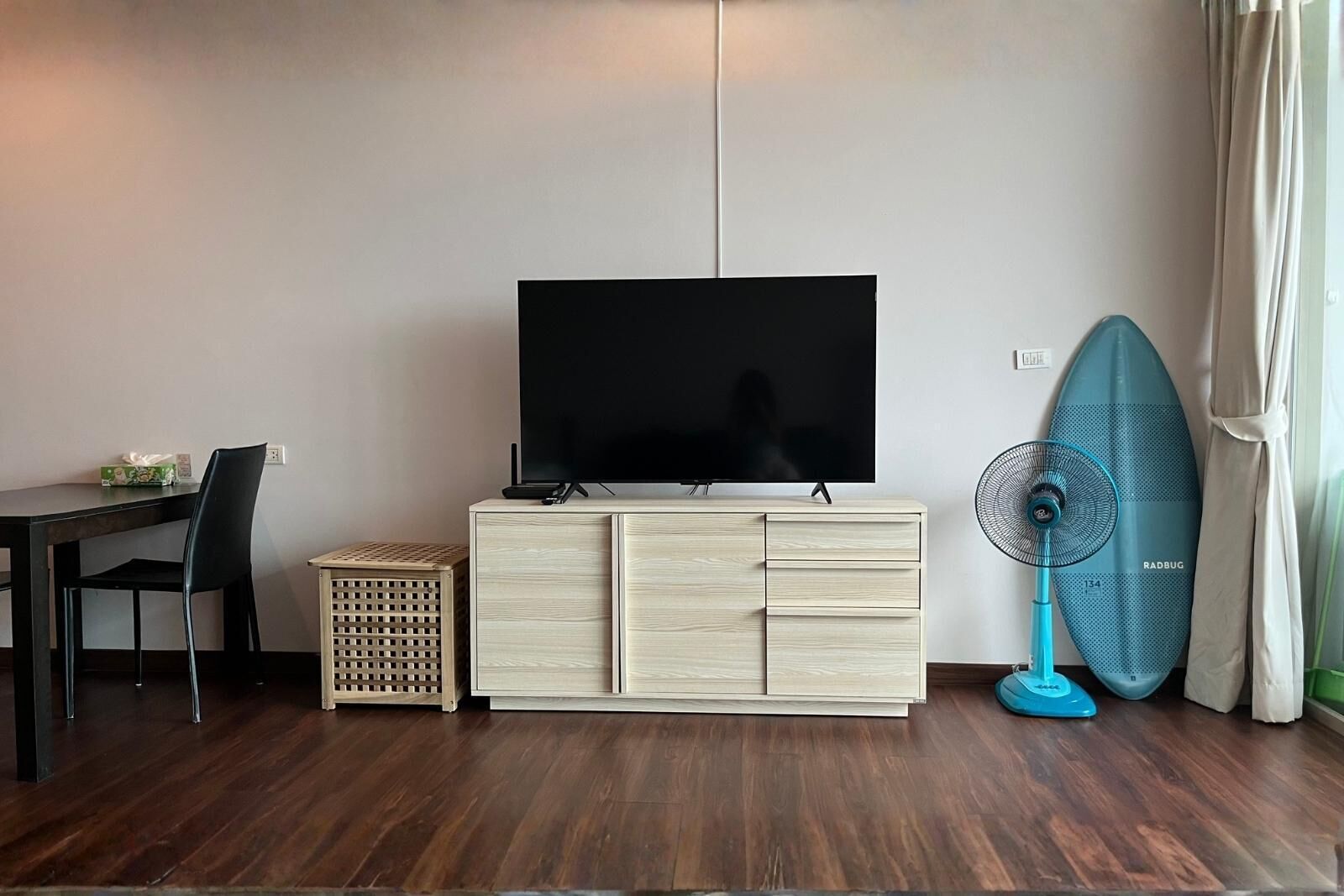 Una habitación con una televisión sobre un mueble de madera, una mesa de comedor con sillas, un ventilador azul y una ventana con cortinas.