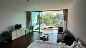 Camera d'albergo moderna con un grande letto, un divano, una scrivania con televisore e un balcone vista verde.