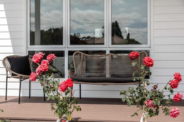 Relax in a serene outdoor seating area with a wicker chair and bench, surrounded by lush greenery and vibrant flowers.