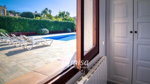 A view from a window to a swimming pool with lounge chairs, a hedge, and a white door.