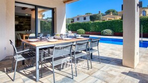 An outdoor patio with a wooden table and chairs, a swimming pool in the background, and a view of the surrounding area.