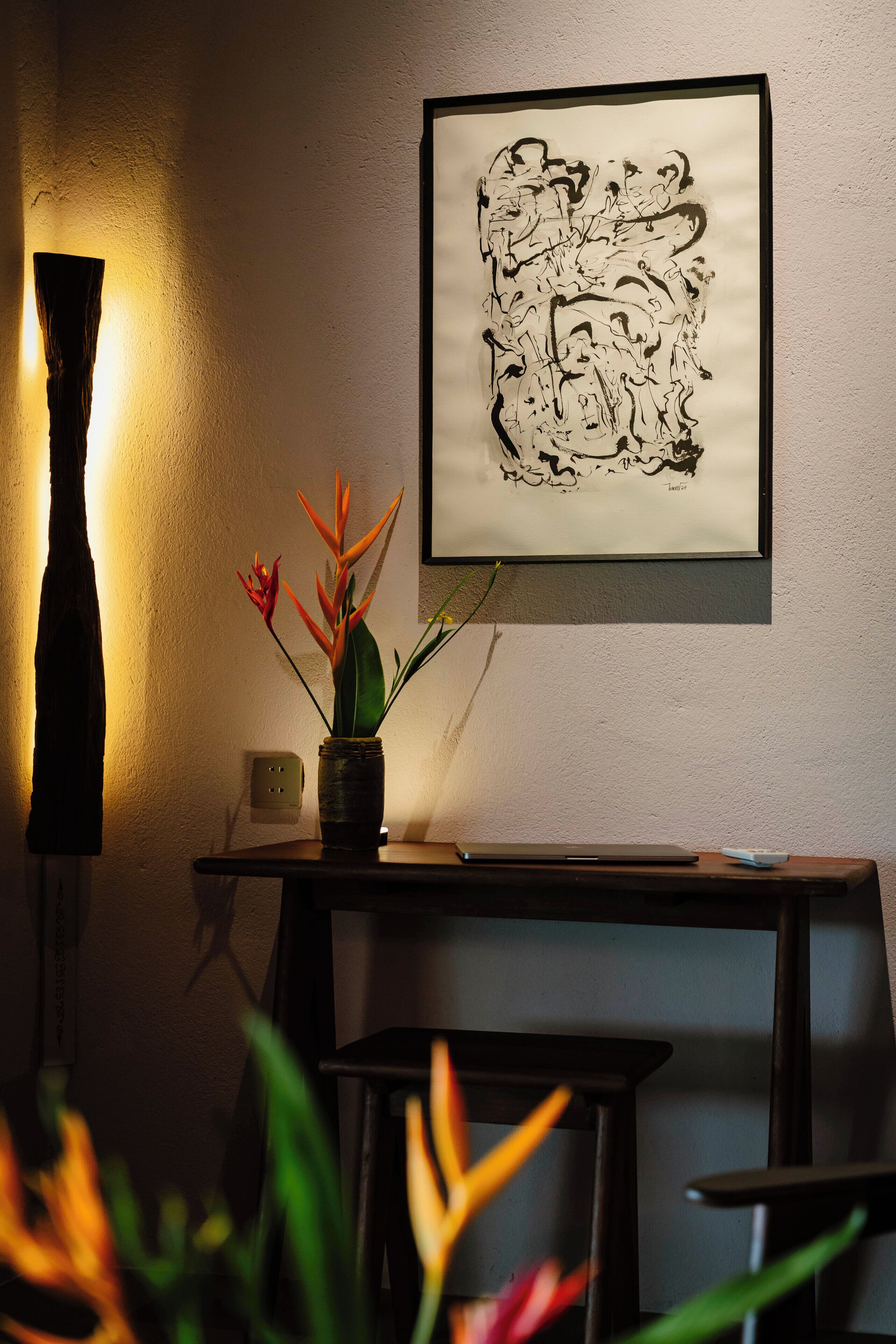 A room with a desk, a vase with flowers, a framed abstract artwork, and a lamp.