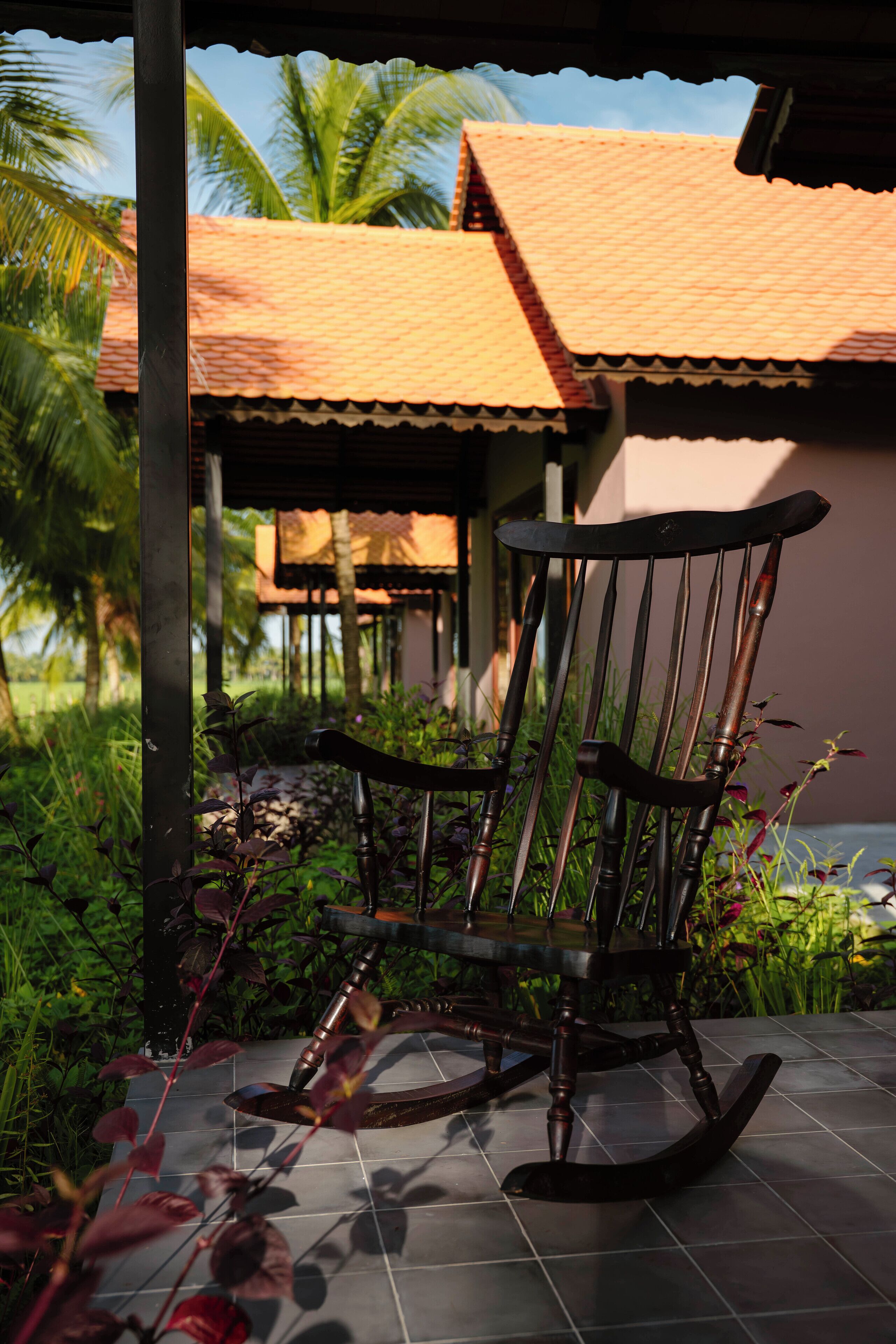 A rocking chair on a tiled patio with a view of a building with a tiled roof and palm trees.