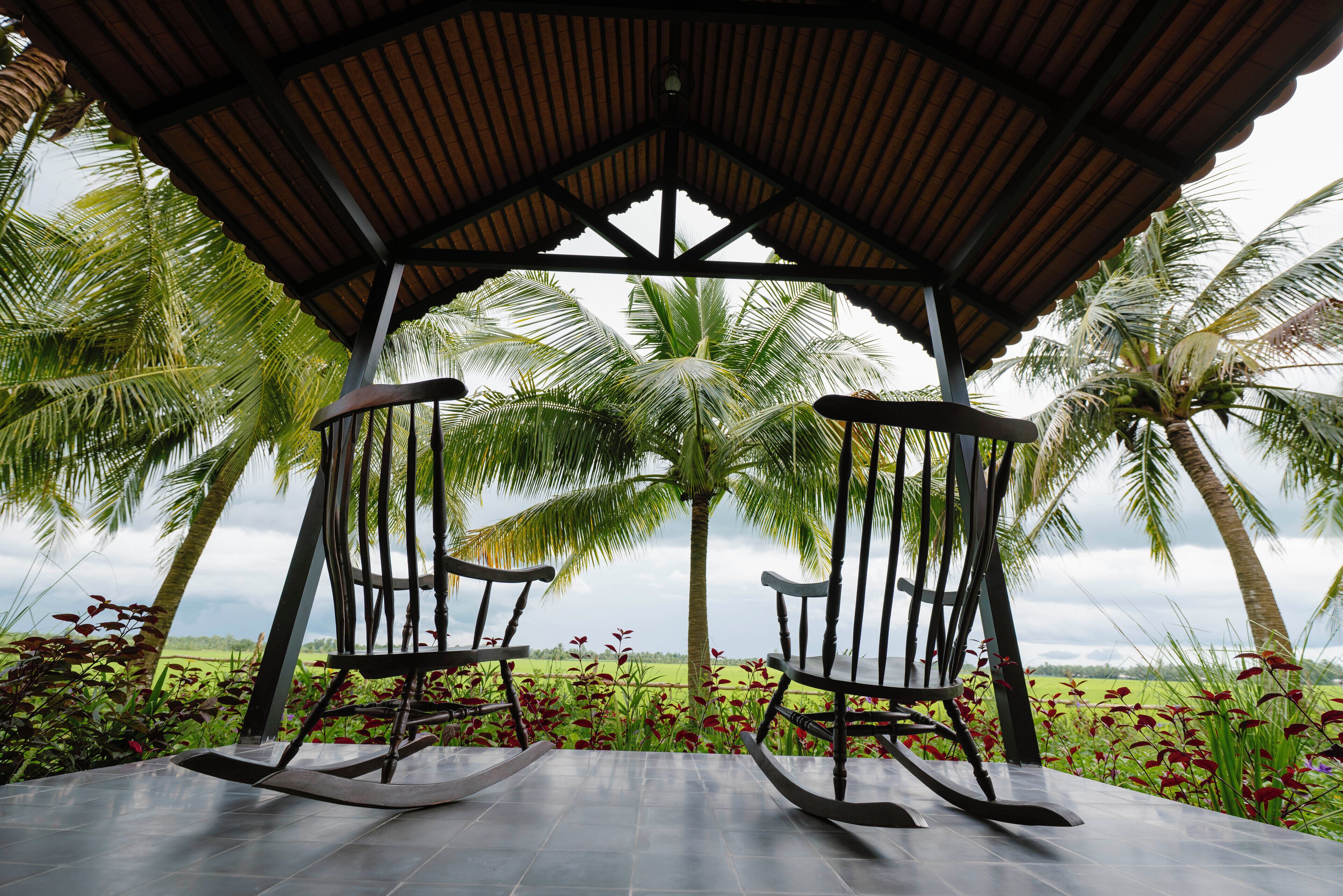 A gazebo with a tiled floor and two rocking chairs, surrounded by palm trees and a view of a green field.