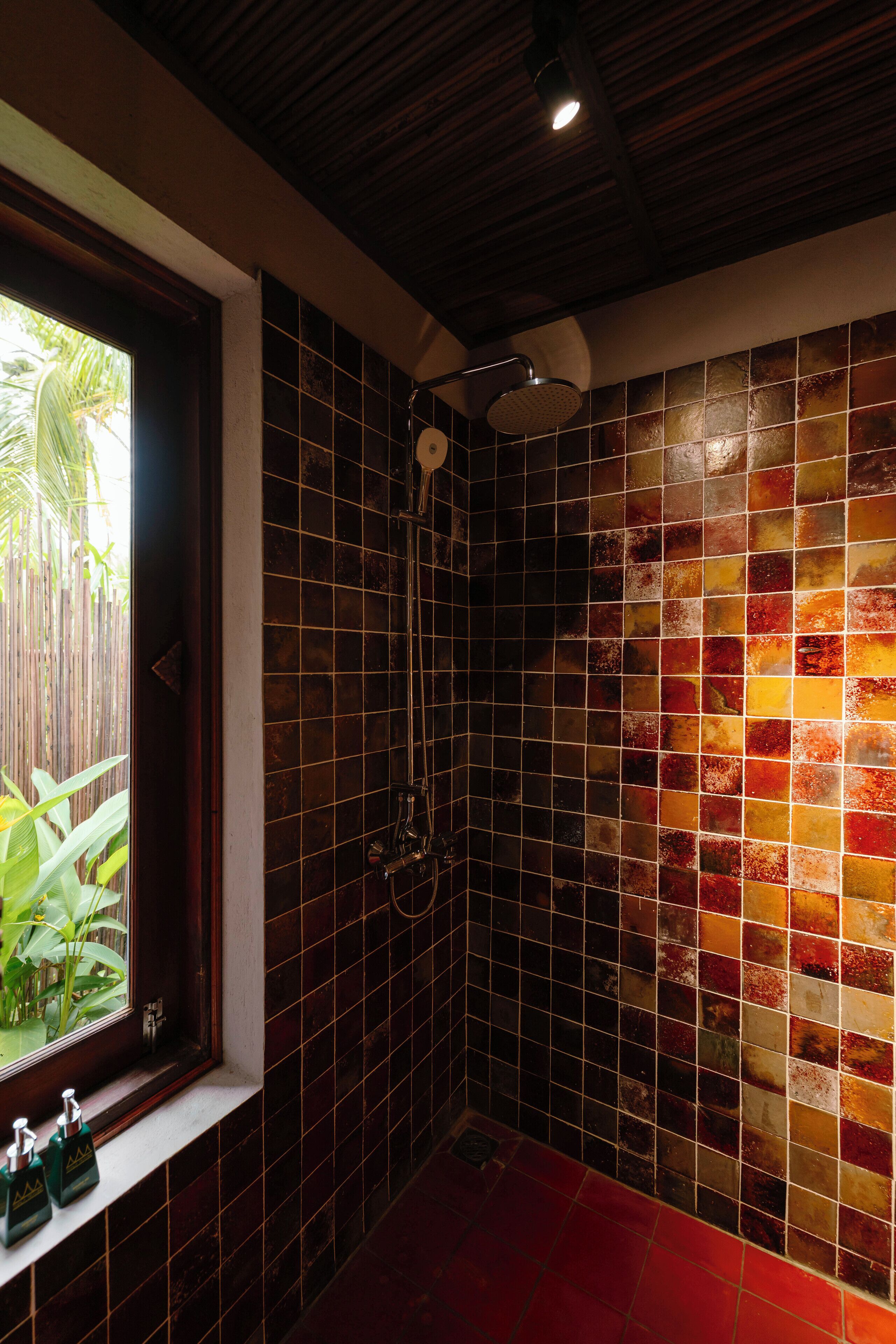 A bathroom with a shower, tiled walls, a window with plants, and a red floor.