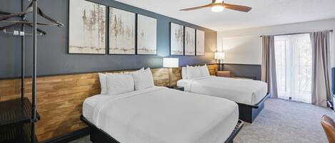 A hotel room with two beds, a ceiling fan, artwork on the wall, and a window with curtains.