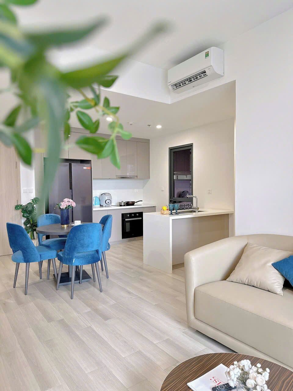 Una sala moderna con un sofá color beige, una mesa redonda y un comedor con sillas azules y un jarrón con flores.