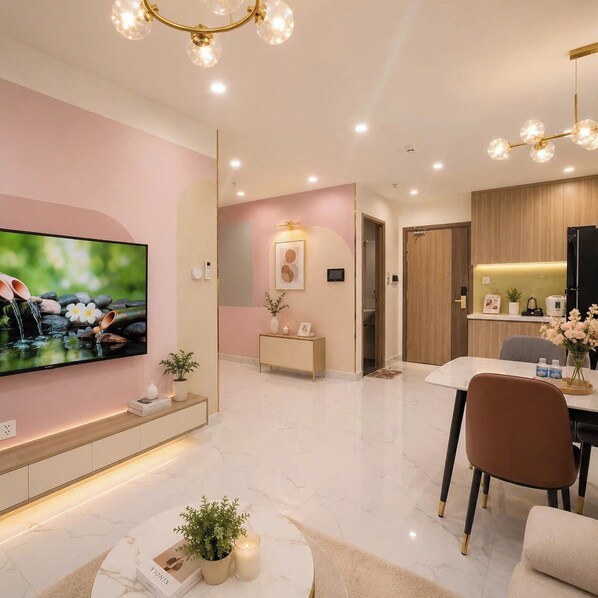 A modern living room with a flat-screen TV, a dining area with a table and chairs, and a kitchen area with a refrigerator and countertop. - Lyshing Apartment in Vinhomes Grand Park (Ho Chi Minh City)