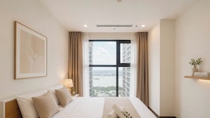 A hotel room with a large bed, a view of a cityscape, a framed artwork on the wall, and a bedside table with a lamp. - Lyshing Apartment in Vinhomes Grand Park (Ho Chi Minh City)