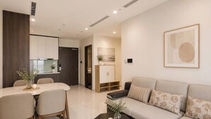 A modern living room with a sofa, a dining table with chairs, a coffee table, and a kitchen area in the background. - Lyshing Apartment in Vinhomes Grand Park (Ho Chi Minh City)