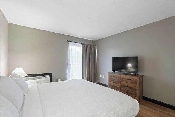 Une chambre d’hôtel avec un grand lit, un téléviseur posé sur une commode en bois, une fenêtre avec des rideaux et un climatiseur fixé au mur.