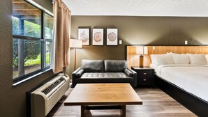 A hotel room with a large bed, a sofa, a table, and a window with curtains. There are three wall-mounted pictures and a heater on the wall. - Renovated 1 King Pet Friendly (Townsend)
