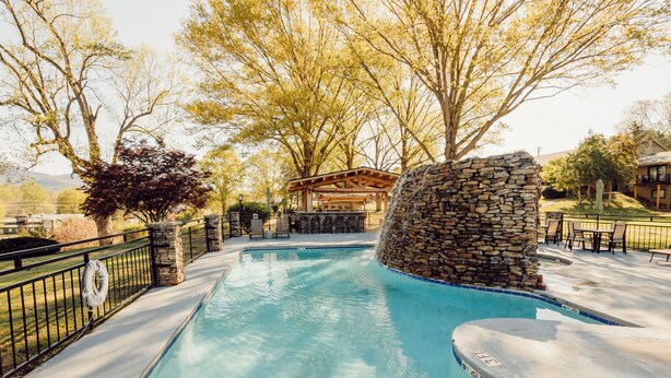 A swimming pool with a stone wall, surrounded by a fence, and a covered seating area in the background. - Renovated 1 King Pet Friendly (Townsend)