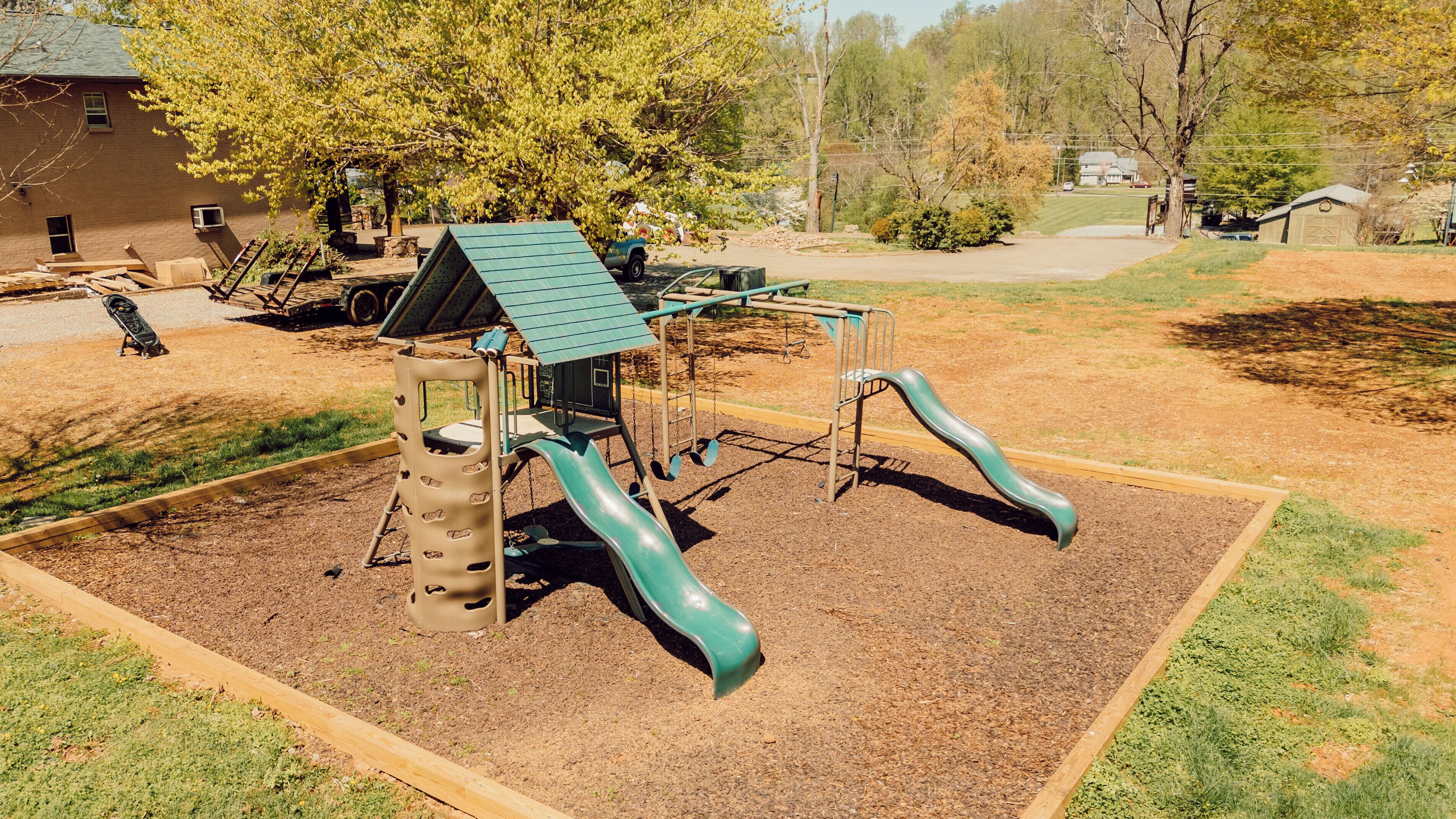 A playground with a slide and climbing structure, surrounded by a fenced area with grass and trees.