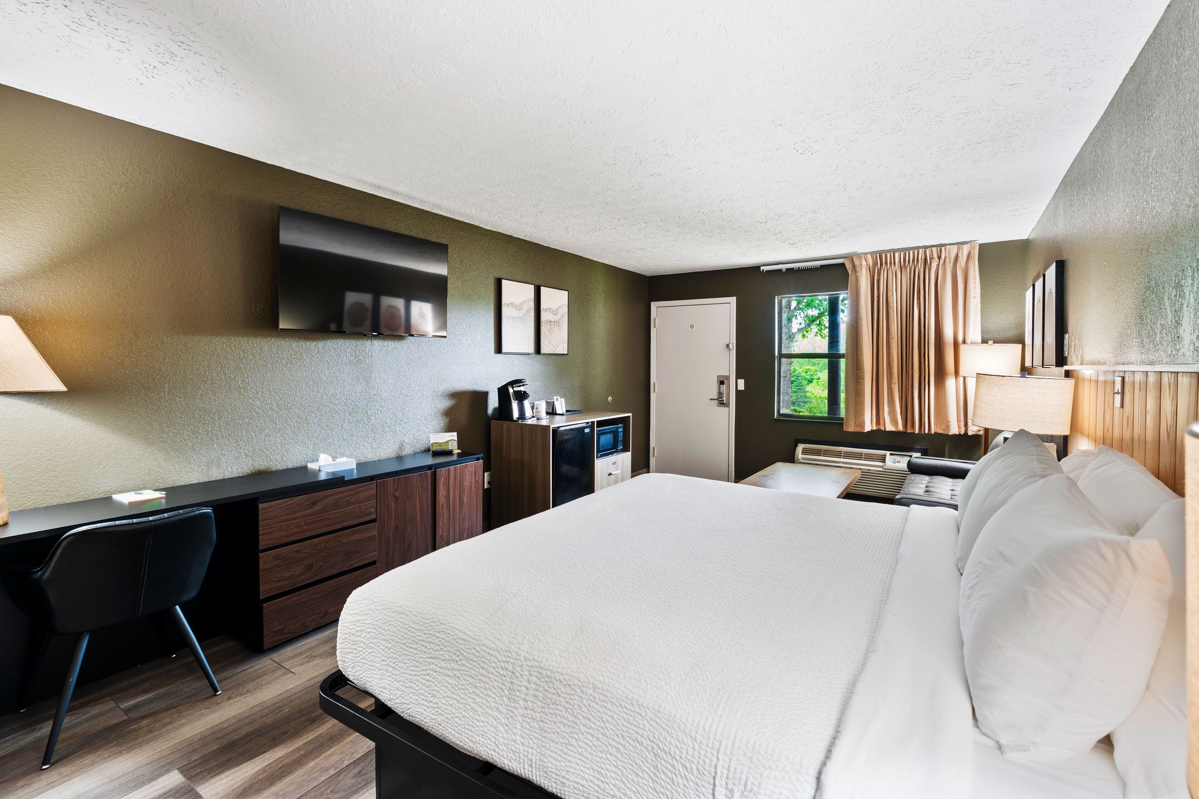A hotel room with a large bed, a desk with a chair, a small kitchenette, and a window with curtains.