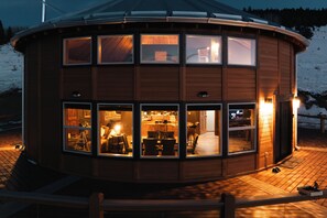 A wooden cabin with multiple windows, a kitchen, and a dining area visible through the windows.