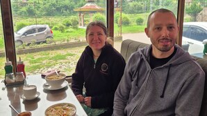 Due persone sedute a un tavolo con vista su un paesaggio rigoglioso, che include un gazebo e una bandiera.