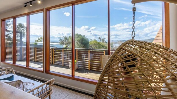 Une pièce dotée de grandes fenêtres, d’un fauteuil en osier et offrant une vue sur un balcon avec une balustrade.